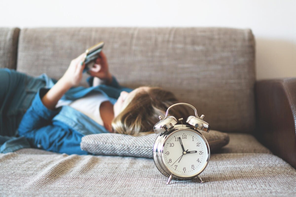Girl on phone whilst lying down on sofa with an alarm clock showing the time