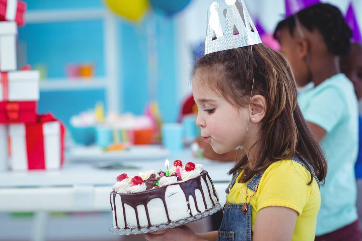 Young girl in dungarees blowing out candles on a birthday cake with cherries on top