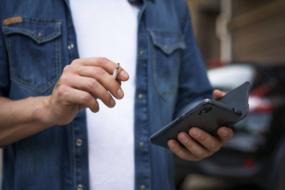close up of man's hands holding smartphone in left hand and lit cigarette in right hand.