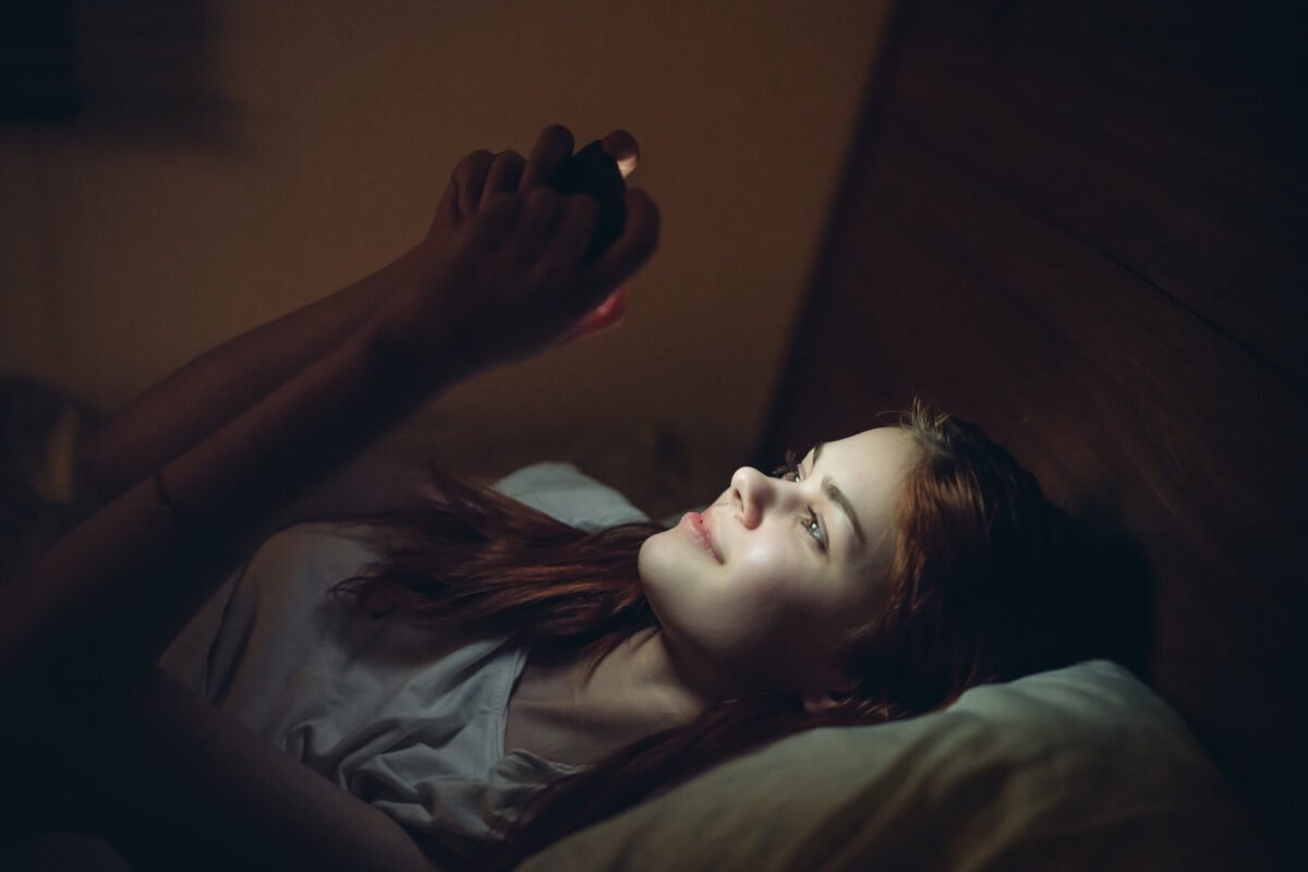 teenage girl on phone lying in bed in dark while phone lights her face