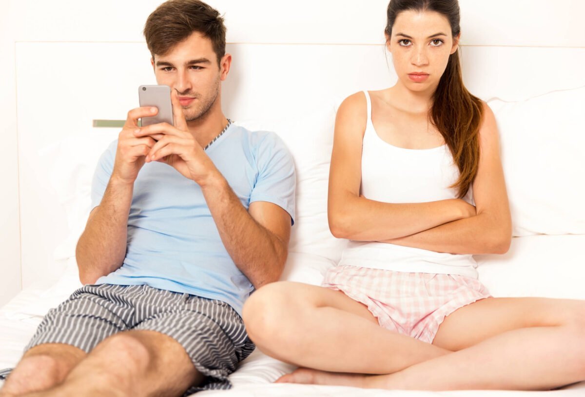 Man and woman sat on bed in pyjamas, man is doomscrolling on phone while woman looks unhappy with arms and legs crossed.