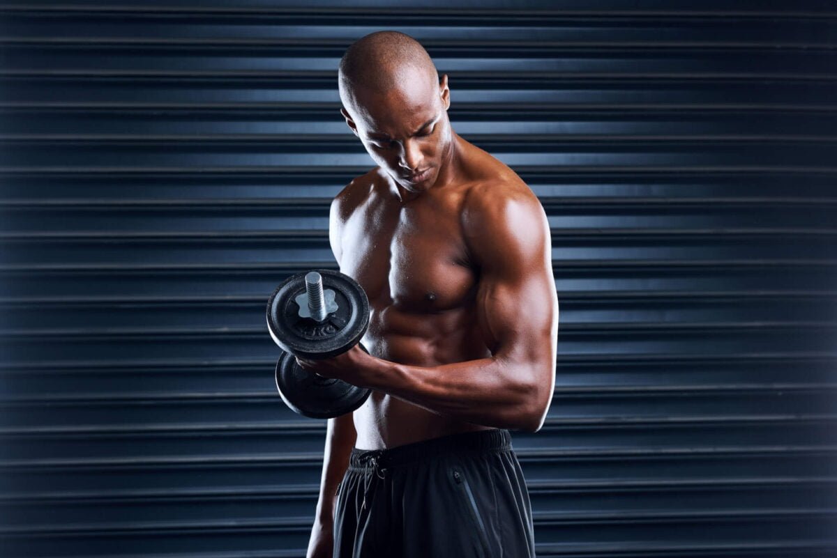 topless black muscular man working out with dumbell in front of metal shutter
