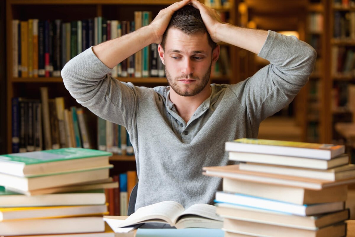 young man with hands on head surrounded by books in library looking stressed
