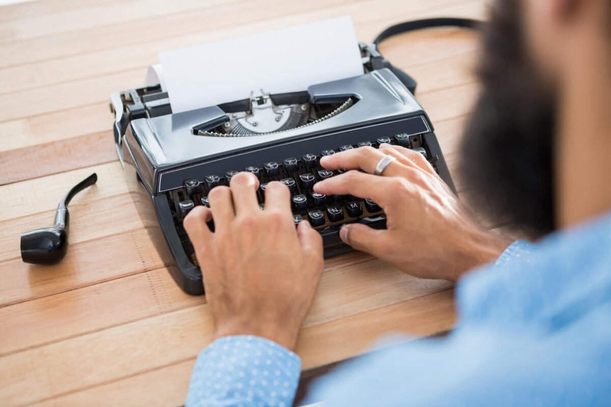 Man with beard typing at a typewriter with a smoking pipe on the desk