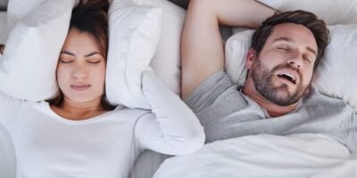 Woman covering ears with pillow while man is snoring next to her in white bed - he has sleep apnea and adhd