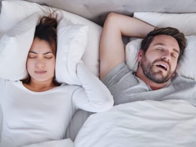 Woman covering ears with pillow while man is snoring next to her in white bed - he has sleep apnea and adhd