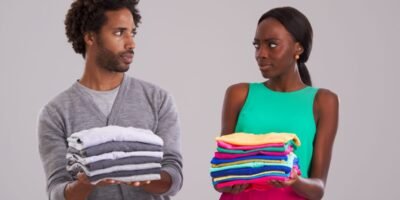 People, studio and pile of laundry in hand for neat, cleaning day or washing clothes together. Couple, surprise and shocked for organised wardrobe with color choice, fashion, perfectionism or ocd on grey background