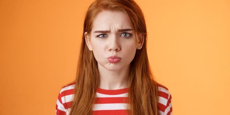 Childish offended cute redhead girl pouting, sulking upset, frowning on orange background