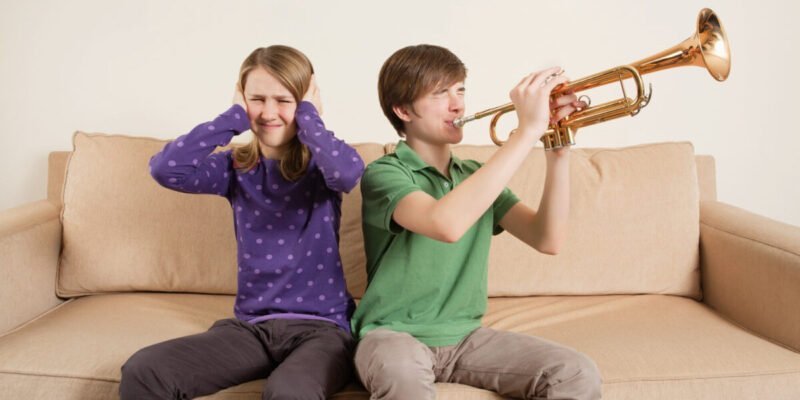 teenage boy and girl on sofa. boy is playing trumpet and girl has sensory processing disorder with hands over ears